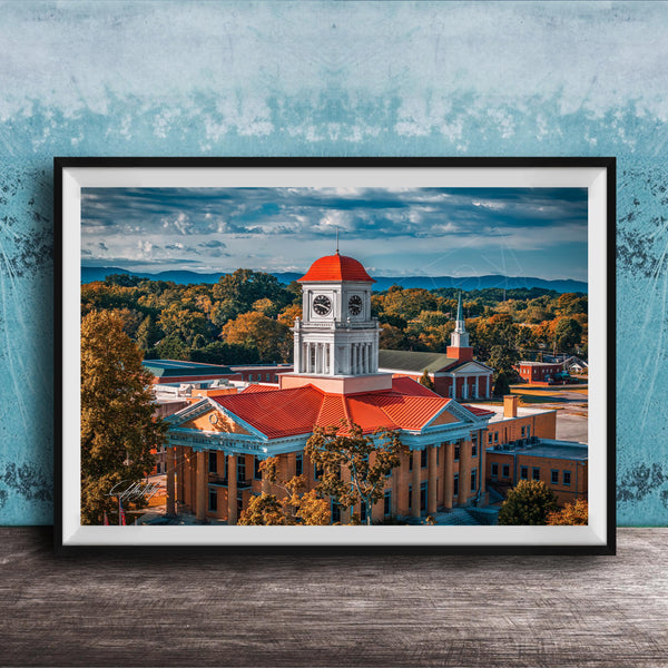 A framed photograph of a historic building with a clock tower, surrounded by trees and mountains in the background.