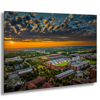 The image is an aerial view of a large sports stadium surrounded by a cityscape, with a dramatic sky in the background.