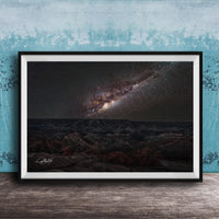 The image is a framed photograph of a night sky with a galaxy, placed on a wooden surface against a blue wall.
