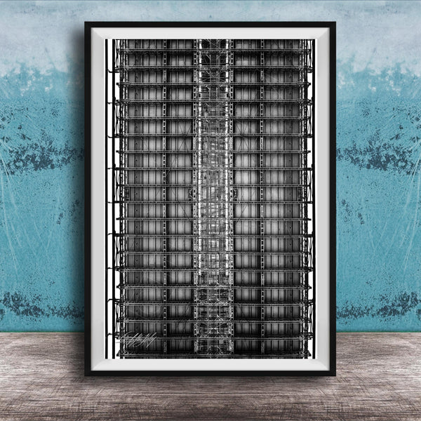 The image is a black and white photograph of a tall, narrow building with a grid-like pattern of windows and steel beams. The photograph is framed and displayed on a wooden surface against a blue wall.