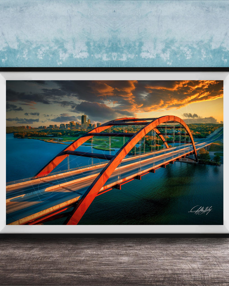 The image is a framed photograph of a red arch bridge spanning across a body of water, with a city skyline visible in the background.