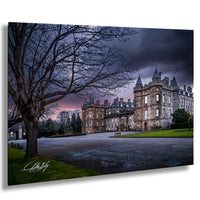 A large, historic castle with multiple towers and turrets, surrounded by a well-manicured lawn and trees, set against a dramatic sky with a pink and purple sunset.