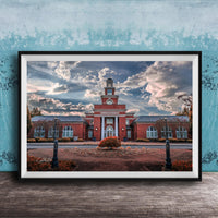 A framed photograph of a red brick building with a clock tower, surrounded by a brick walkway and landscaping, set against a cloudy sky.