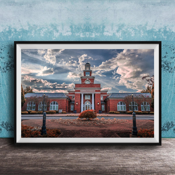 A framed photograph of a red brick building with a clock tower, surrounded by a brick walkway and landscaping, set against a cloudy sky.
