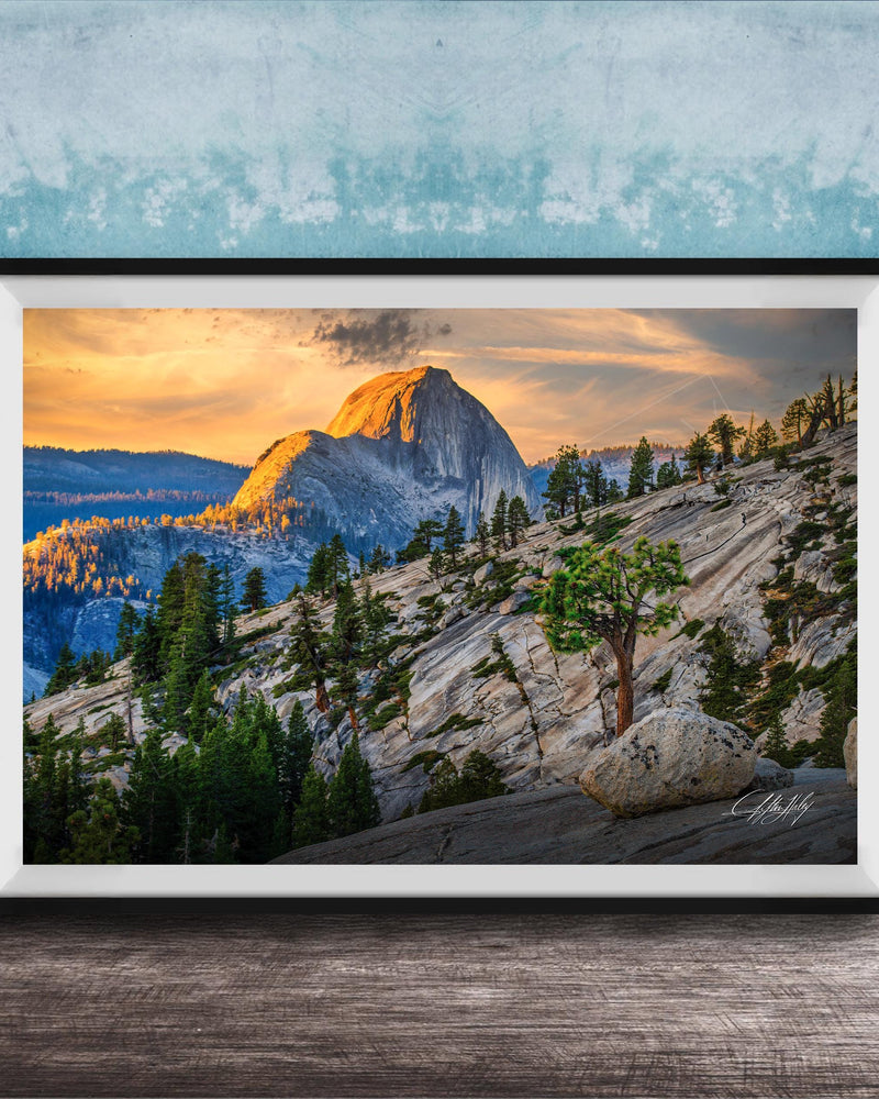 A framed photograph depicting a mountainous landscape with a large, snow-capped peak in the distance, surrounded by trees and rocky terrain. The sky is filled with warm, golden light, creating a serene and picturesque scene.