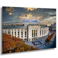 the Yankee Stadium, a large sports stadium located in New York City. The stadium is surrounded by trees with autumn foliage, and the sky is filled with clouds. The stadium is prominently displayed with the words "Yankees Stadium" written on it.