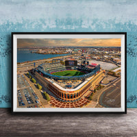 An aerial view of a baseball stadium with a large field, surrounding parking lots, and a body of water in the background.