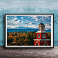 A framed photograph of a red brick building with a dome, set against a backdrop of a blue sky with clouds and a mountainous landscape.