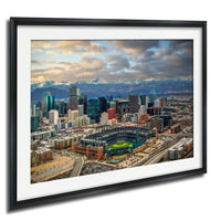The image is a framed aerial view of a large city with a baseball stadium in the center, surrounded by tall buildings and mountains in the background.