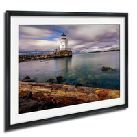 A framed photograph depicting a lighthouse on a rocky shore, with a calm body of water in the foreground and a cloudy sky above.