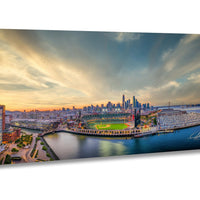 A panoramic view of a city skyline at sunset, with a baseball field and a body of water in the foreground.