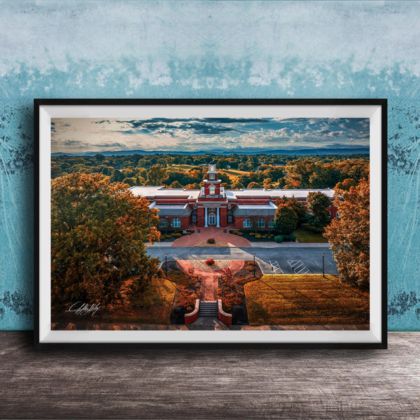 A framed aerial view of a red brick building with a clock tower, surrounded by trees with autumn foliage, set against a blue sky with clouds.