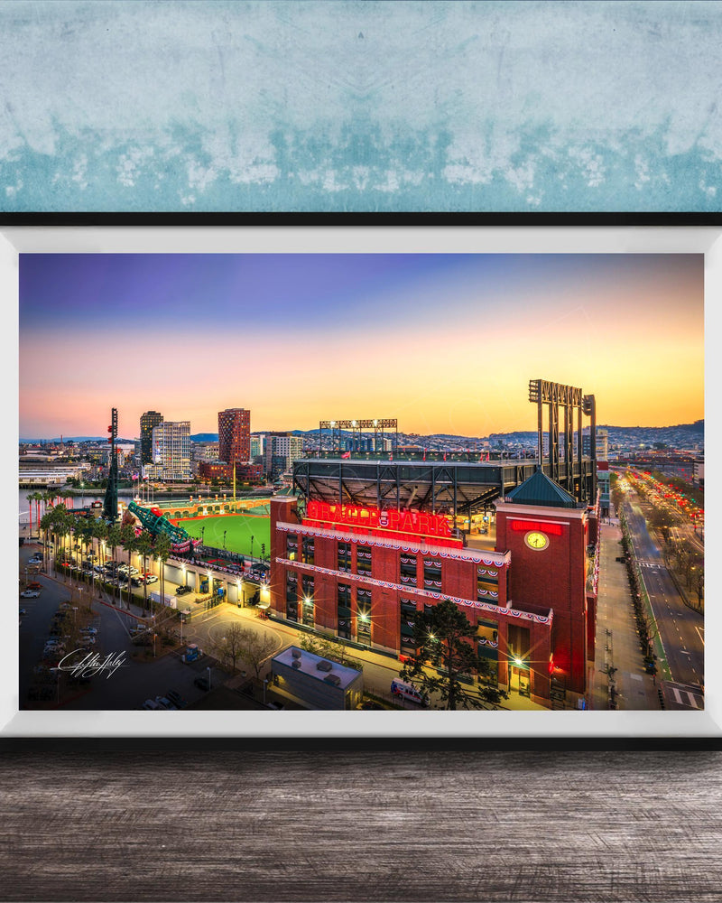 A framed photograph of a city skyline at dusk, with a large stadium and surrounding buildings illuminated against a colorful sunset sky.