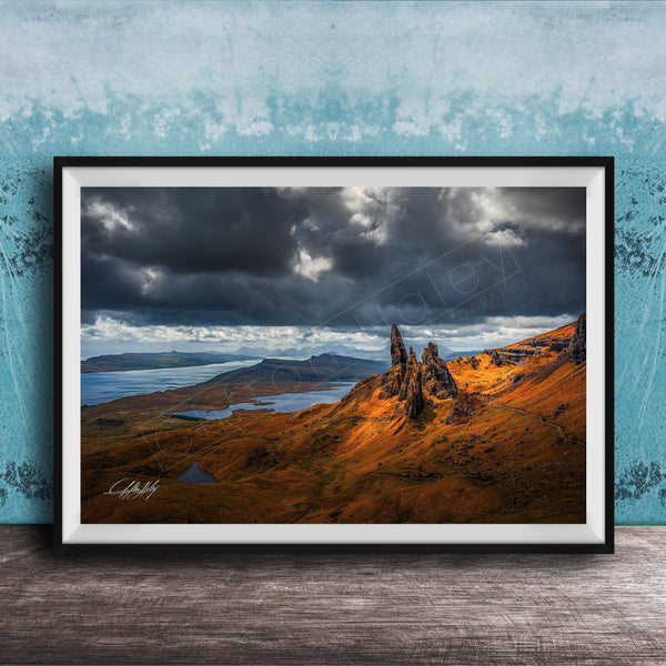 The image is a framed photograph depicting a dramatic landscape with a rocky outcrop, a lake, and a cloudy sky.