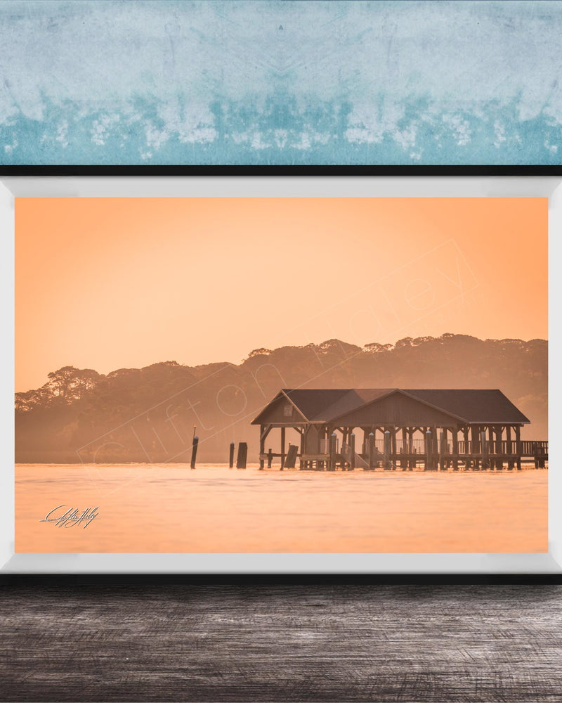 A framed photograph of a wooden pier extending into a body of water, with a warm orange and yellow sunset sky in the background.
