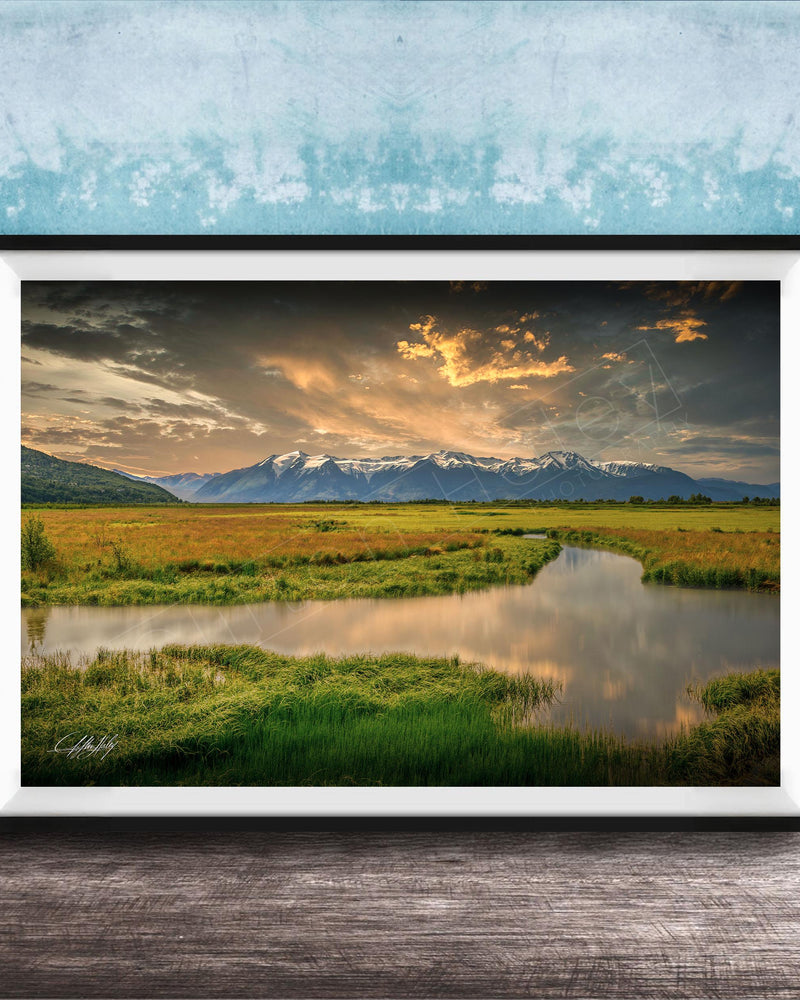 The image is a framed landscape photograph depicting a serene scene of a river flowing through a grassy field, with mountains in the background under a dramatic sky.