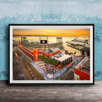 The image is a framed aerial view of a cityscape with a large stadium, buildings, and a body of water in the background.