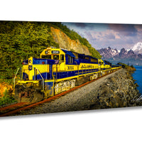 A yellow and blue Alaska train is traveling along a track next to a body of water, with mountains in the background.