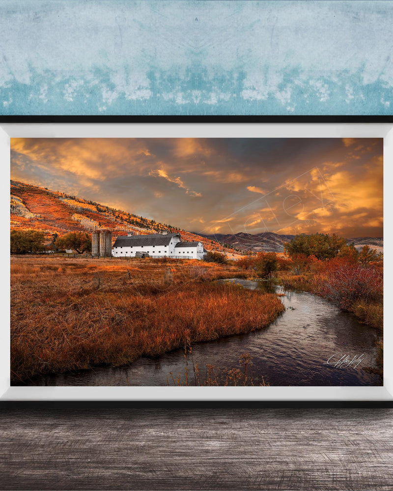 a picturesque rural landscape with a farmhouse nestled among rolling hills and a winding stream. The sky is filled with vibrant orange and yellow hues, creating a warm and serene atmosphere. The overall scene is a beautiful representation of nature and the tranquility of rural life.