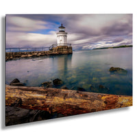 A white lighthouse stands on a rocky shore, overlooking a calm body of water with a cloudy sky in the background.