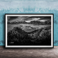 A black and white landscape photograph depicting a mountainous, forested area with a winding river or stream in the foreground.