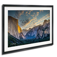 a breathtaking view of the majestic Yosemite Falls, with its cascading waters flowing down the towering granite cliffs. The sky above is a beautiful canvas of blue and orange hues, creating a stunning backdrop for the natural landscape. The photograph is framed and hangs on a wall, capturing the essence of this awe-inspiring scene.