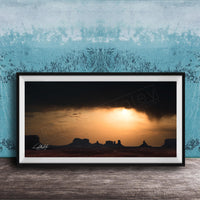 a framed photograph of a desert landscape at sunset, with silhouetted mountains in the background and a dramatic sky filled with clouds and the sun setting behind them.