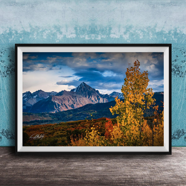 The image is a framed photograph depicting a scenic mountain landscape with a mix of autumnal and winter colors, including vibrant yellow and orange foliage in the foreground and majestic snow-capped peaks in the background.