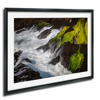 A framed photograph depicting a rocky coastline with flowing water and patches of green moss.