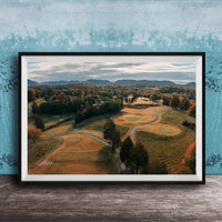 The image is a framed aerial view of a golf course surrounded by trees and rolling hills, with a blue sky and clouds overhead.