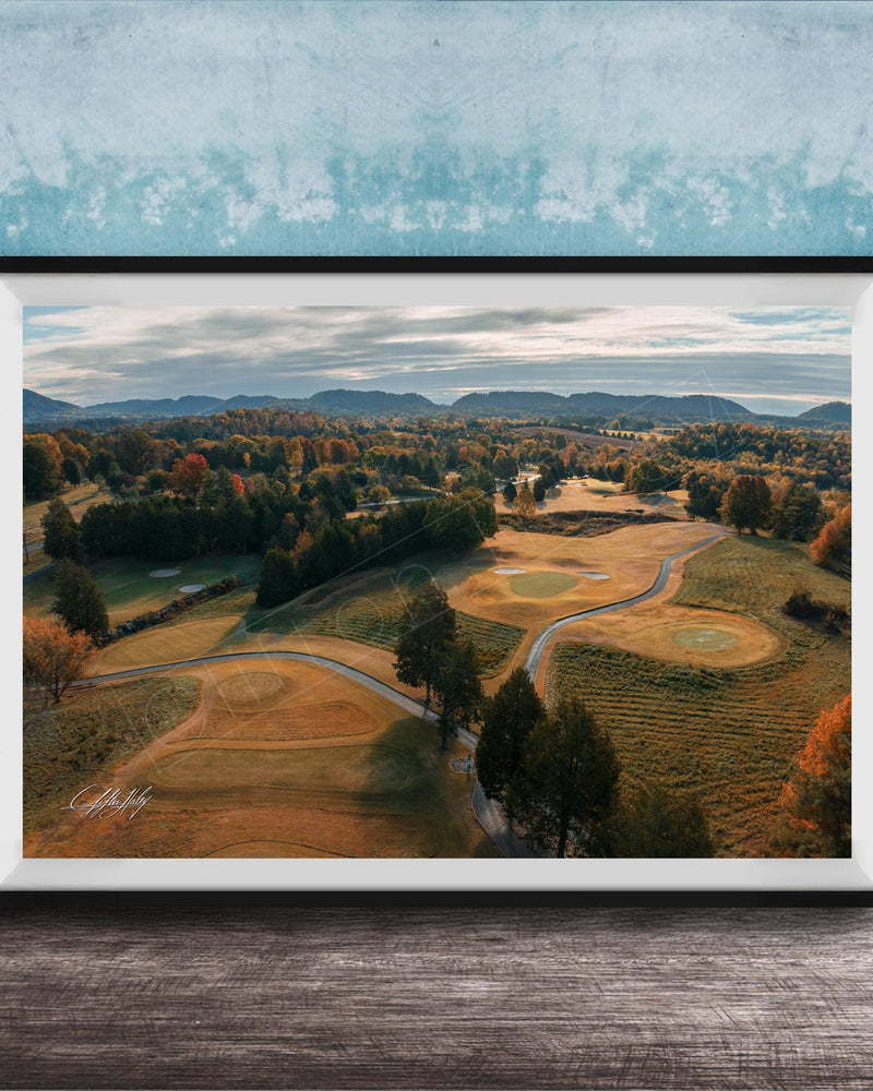 The image is a framed aerial view of a golf course surrounded by trees and rolling hills, with a blue sky and clouds overhead.