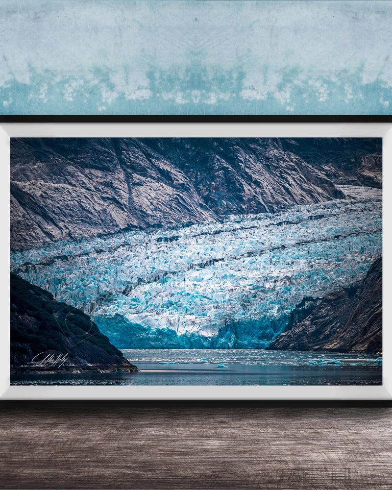 A framed photograph of a large, majestic glacier with a blue-green hue, set against a backdrop of rugged mountains and a serene body of water.