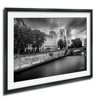A black and white photograph of the Notre-Dame Cathedral in Paris, France, with a river running alongside the cathedral and surrounding buildings.