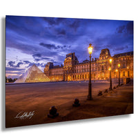 the Louvre Museum in Paris, France, with its iconic glass pyramid and illuminated architecture against a dark blue sky.