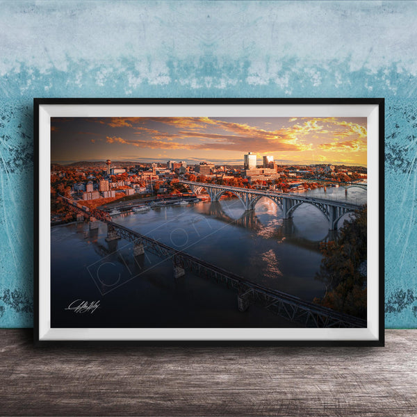 The image is a framed photograph depicting a cityscape with a bridge spanning a river, set against a backdrop of a colorful sunset sky.