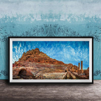 The image is a framed photograph of a desert landscape with a rocky hillside, a cave, and a few buildings or structures in the background.