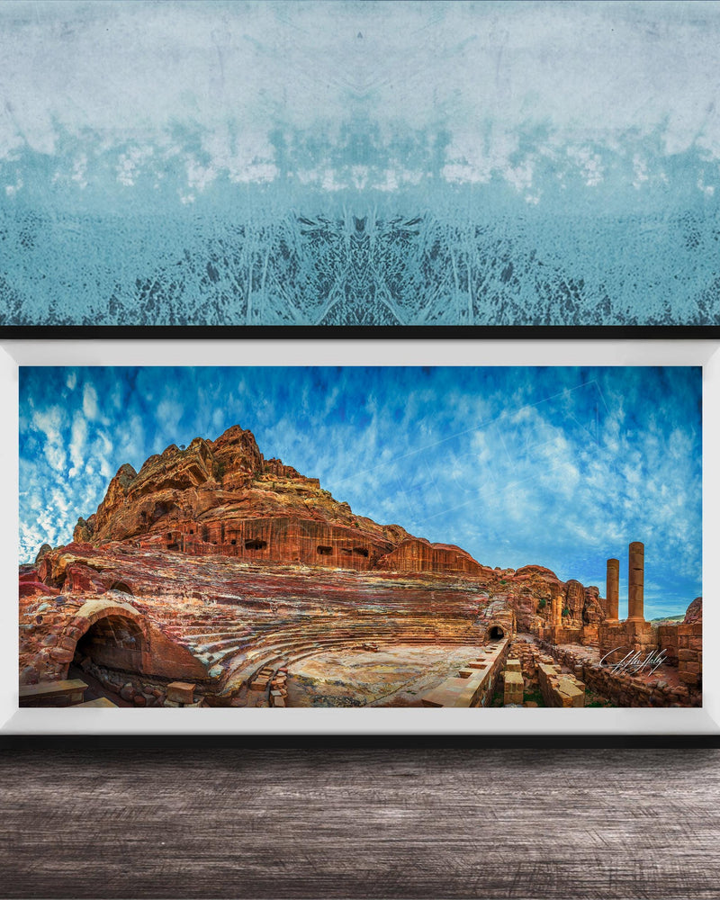 The image is a framed photograph of a desert landscape with a rocky hillside, a cave, and a few buildings or structures in the background.