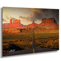 a long, straight road cutting through a vast, arid landscape with towering red rock formations in the background. The sky above is filled with clouds, creating a dramatic atmosphere. The road appears to be the main focus of the image, leading the viewer's eye towards the distant mountains.