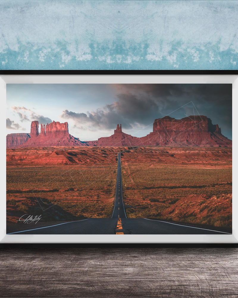 The image is a framed photograph depicting a desert landscape with a winding road leading towards a series of red rock formations in the distance.