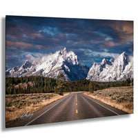 A scenic mountain road winding through a forested landscape with snow-capped peaks in the background.