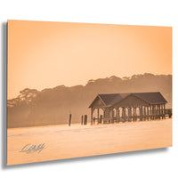A wooden pier extends into a body of water, with a small building on the pier and a line of trees in the background. The sky is hazy, creating a warm, golden glow over the scene.
