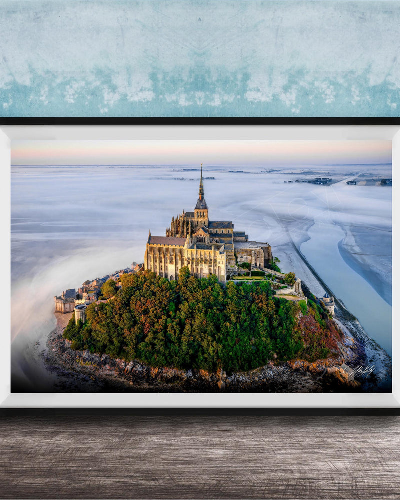 A framed photograph of the iconic Mont Saint-Michel island in Normandy, France, with its distinctive castle perched on a rocky outcrop surrounded by lush greenery.