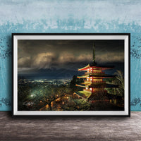 A framed photograph of a traditional Japanese pagoda, illuminated at night against a backdrop of a mountainous landscape and a cloudy sky.