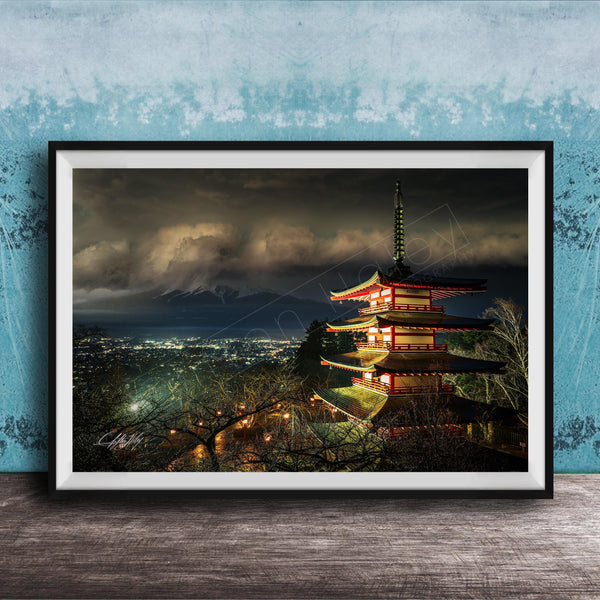 A framed photograph of a traditional Japanese pagoda, illuminated at night against a backdrop of a mountainous landscape and a cloudy sky.