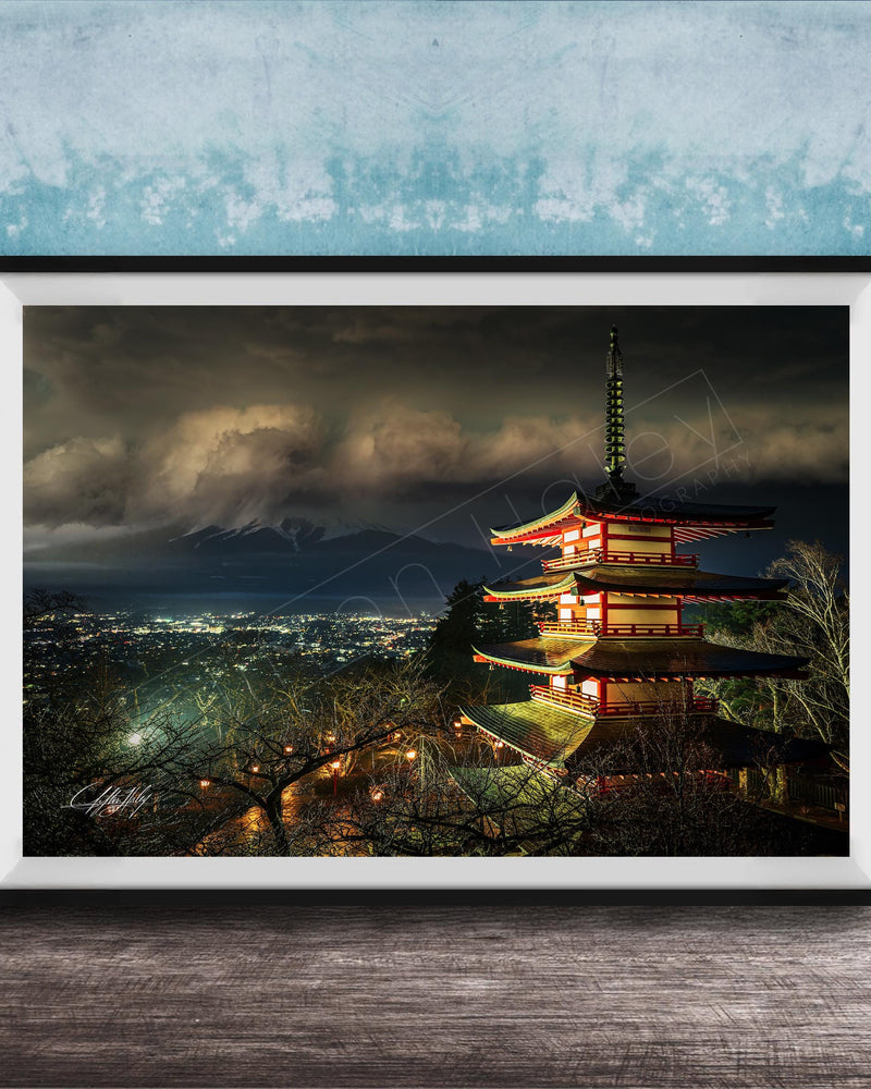 A framed photograph of a traditional Japanese pagoda, illuminated at night against a backdrop of a mountainous landscape and a cloudy sky.