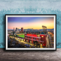 A framed photograph of a city skyline at dusk, with a large stadium and surrounding buildings illuminated against a colorful sunset sky.