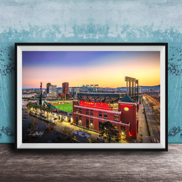 A framed photograph of a city skyline at dusk, with a large stadium and surrounding buildings illuminated against a colorful sunset sky.