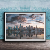 A framed photograph of a city skyline at sunset, with the buildings reflected in the water below.