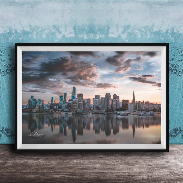 A framed photograph of a city skyline at sunset, with the buildings reflected in the water below.