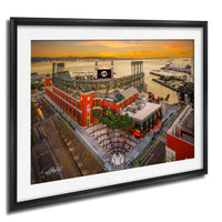 The image is a framed aerial view of a large stadium with a red and white exterior, surrounded by palm trees and other buildings. The stadium appears to be a sports venue, with a large screen visible in the background. The sky is filled with warm, orange and yellow hues, suggesting a sunset or sunrise. The image is framed with a black border, and the artist's signature is visible in the bottom left corner.